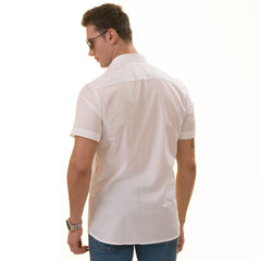 A man wearing a white paisley short sleeve shirt with a spread collar, paired with a red hat and sunglasses, standing against a white background.